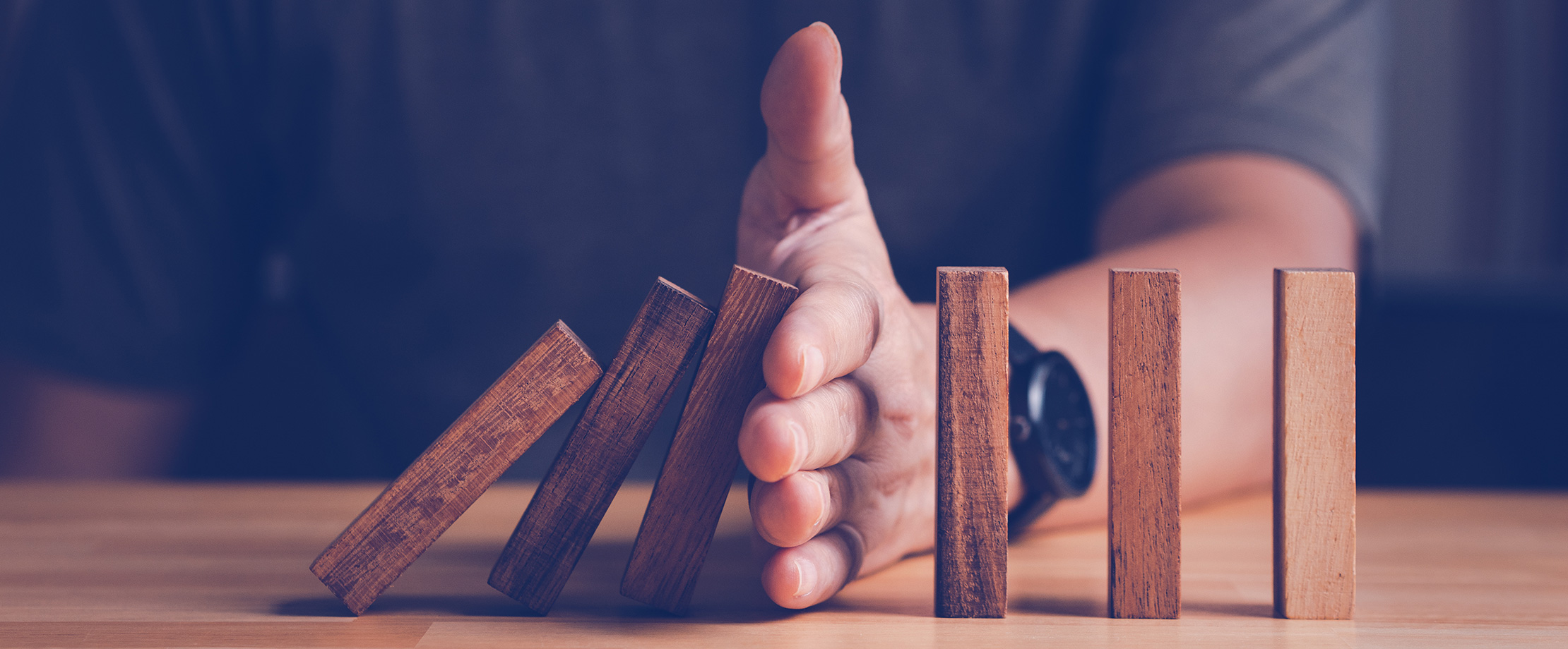 Risiken minimieren Auf einem Tisch sind hölzerne Dominosteine aufgestellt, die umkippen. Ein Mann hält seine Hand zwischen die Steine, um zu verhindern, dass auch die hinteren Steine umfallen.